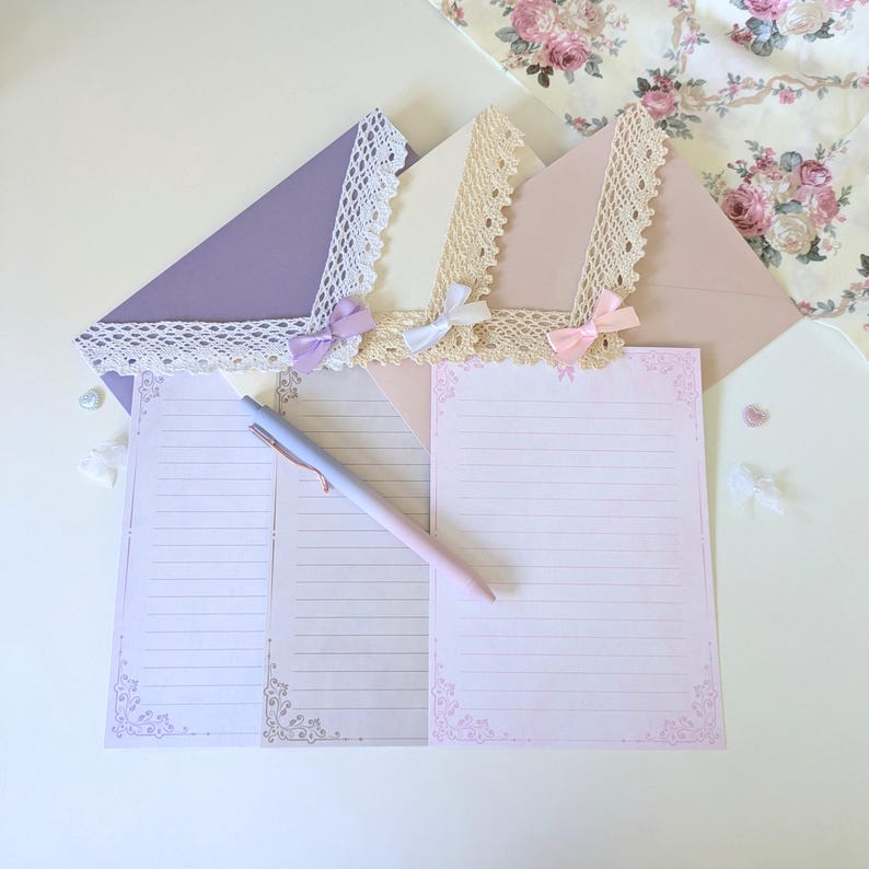 May include: Two sheets of stationery with delicate floral borders and lined paper. The stationery is decorated with lace and pink ribbons. A purple pen is lying on top of the stationery.