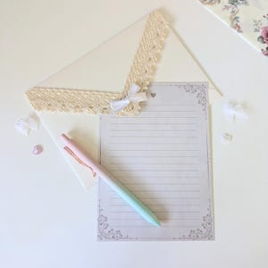 May include: A white envelope with a lace trim and a white bow. A lined sheet of paper with a decorative border is on top of the envelope. A pink and green pen is lying on the paper.