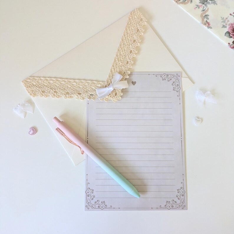 May include: A white envelope with a lace trim and a white bow. A lined sheet of paper with a decorative border is on top of the envelope. A pink and green pen is lying on the paper.