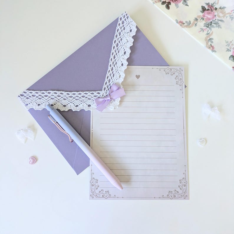 May include: A purple envelope with white lace trim and a lavender bow. A white lined writing paper with a decorative border is on top of the envelope. A light blue and pink pen is lying on the envelope and paper.