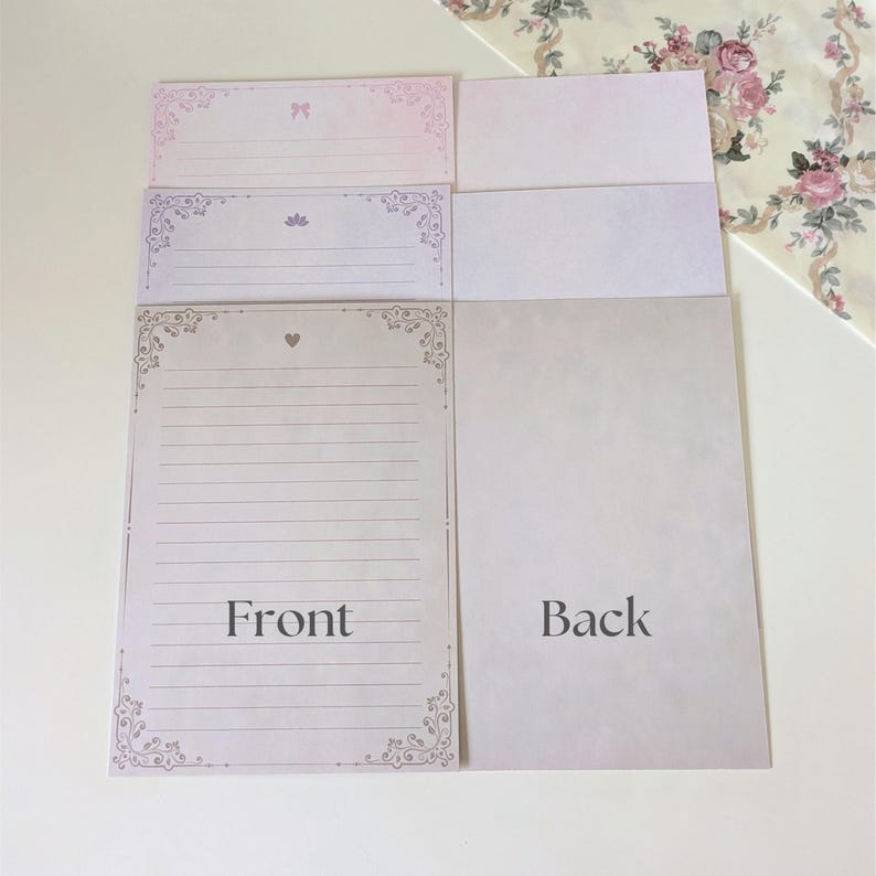 May include: A set of three stationery note cards with decorative borders. The front of the cards feature a pink, purple, and white color scheme with floral designs. The back of the cards are blank. The text "Front" and "Back" is printed on the cards.
