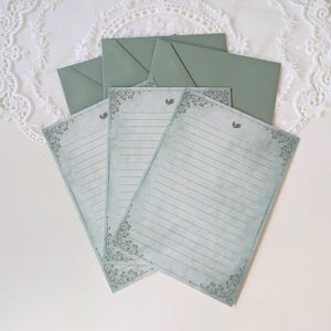 K&ouml;nnte beinhalten: Briefpapier-Set mit linierten Schreibpapieren und passenden Umschl&auml;gen. Das Papier hat eine Vintage-&Auml;sthetik mit dekorativen Eckdesigns und einem verblassten blau-gr&uuml;nen Farbton. Umschl&auml;ge und Papier sind in einem gedeckten Gr&uuml;nton gehalten.