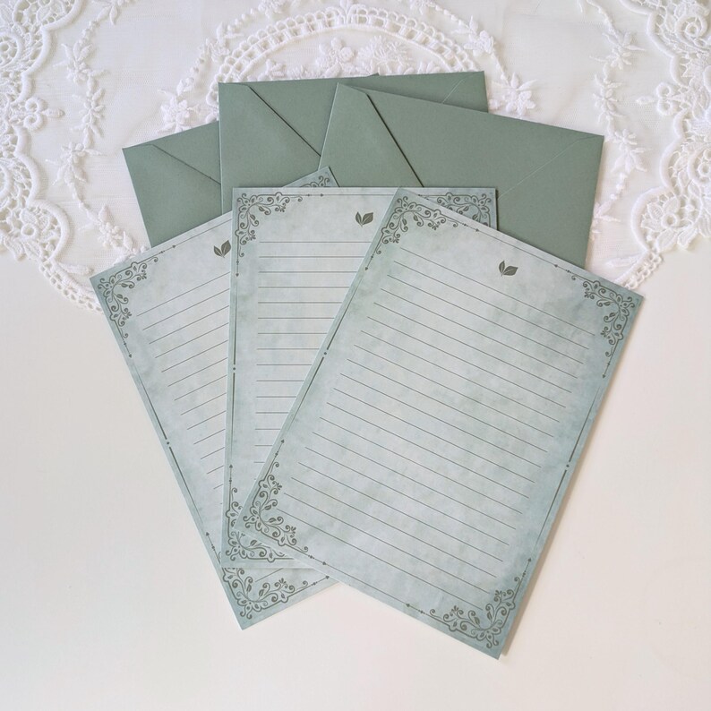 K&ouml;nnte beinhalten: Briefpapier-Set mit linierten Schreibpapieren und passenden Umschl&auml;gen. Das Papier hat eine Vintage-&Auml;sthetik mit dekorativen Eckdesigns und einem verblassten blau-gr&uuml;nen Farbton. Umschl&auml;ge und Papier sind in einem gedeckten Gr&uuml;nton gehalten.