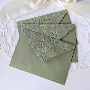 May include: Three sage green envelopes with embossed floral designs on the flaps. The envelopes are arranged on a white lace doily and a cream-coloured satin ribbon. The floral pattern is a repeating design of stylised flowers and leaves.