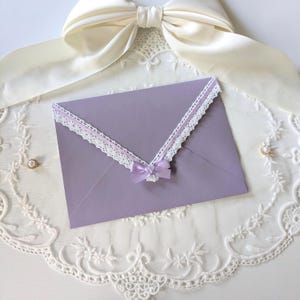 May include: A light purple envelope featuring a white lace trim along the flap edge and a small matching purple satin ribbon bow at the center. The envelope is positioned against a white lace fabric background with a large white satin bow above.