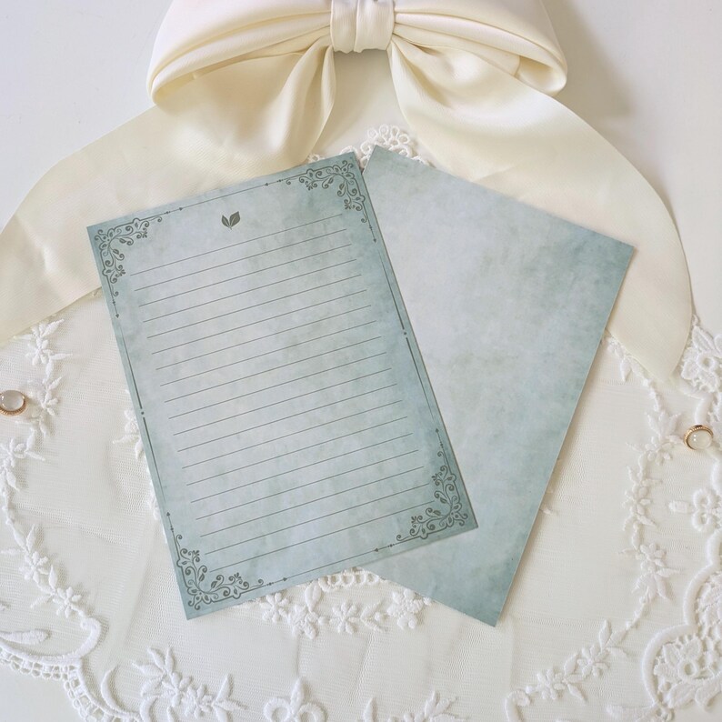 K&ouml;nnte beinhalten: Zwei Briefpapierb&ouml;gen mit Vintage-&Auml;sthetik. Ein Bogen ist mit einem dekorativen Rand und einem kleinen Blattmuster oben versehen. Der andere Bogen hat ein einfarbiges, strukturiertes Aussehen. Das Papier hat einen sanften blau-gr&uuml;nen Farbton.