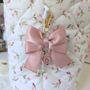 Peut inclure: Un porte-cl&eacute;s en forme de n&oelig;ud en satin rose avec un fermoir dor&eacute; et un petit pendentif en forme de c&oelig;ur avec la lettre "D". Le n&oelig;ud est attach&eacute; &agrave; un tissu matelass&eacute; blanc &agrave; motifs floraux.