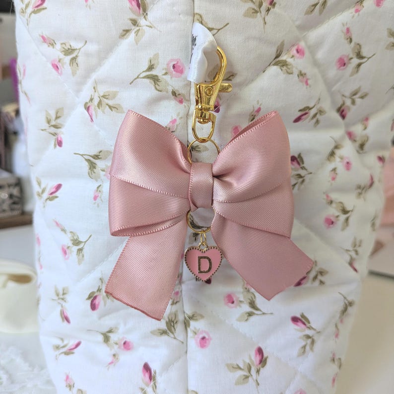 Peut inclure: Un porte-cl&eacute;s en forme de n&oelig;ud en satin rose avec un fermoir dor&eacute; et un petit pendentif en forme de c&oelig;ur avec la lettre "D". Le n&oelig;ud est attach&eacute; &agrave; un tissu matelass&eacute; blanc &agrave; motifs floraux.