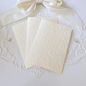 May include: Three cream-colored folded cards featuring an embossed floral vine pattern. The cards are arranged in a fan shape, resting on a white lace doily with a large cream satin ribbon bow positioned at the top center of the composition.