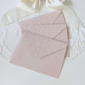 Puede incluir: Sobres de color rosa pálido con patrones florales en relieve. Los sobres están dispuestos sobre una superficie de encaje blanco, con una cinta color crema en el fondo. El diseño es delicado y adecuado para invitaciones o papelería.