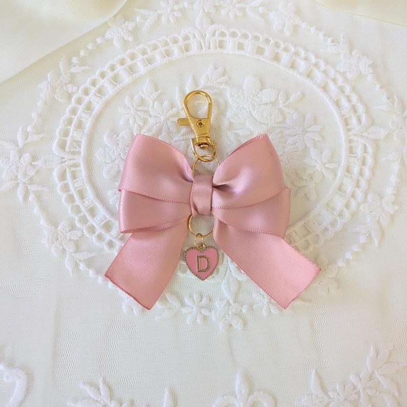 Peut inclure: Un porte-cl&eacute;s en forme de n&oelig;ud en satin rose, avec un fermoir dor&eacute; et un charm en forme de c&oelig;ur. Le charm a un bord rose et la lettre "D" au centre. Le n&oelig;ud est nou&eacute; de mani&egrave;re classique.