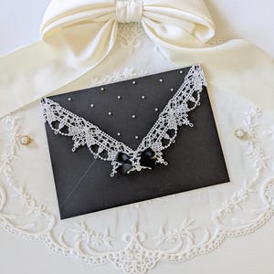 May include: A black envelope featuring a white lace trim along the flap, accented with small white pearls and a central black ribbon bow. The envelope is positioned beneath a large cream-colored satin bow and rests on a white lace doily.
