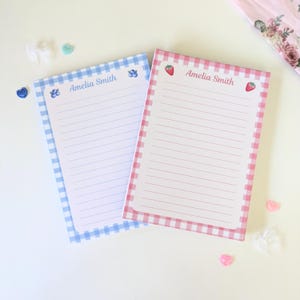 May include: Two personalized notepads with gingham patterns. The blue notepad has blueberries and the pink notepad has strawberries. Both notepads have the name "Amelia Smith" written on them.