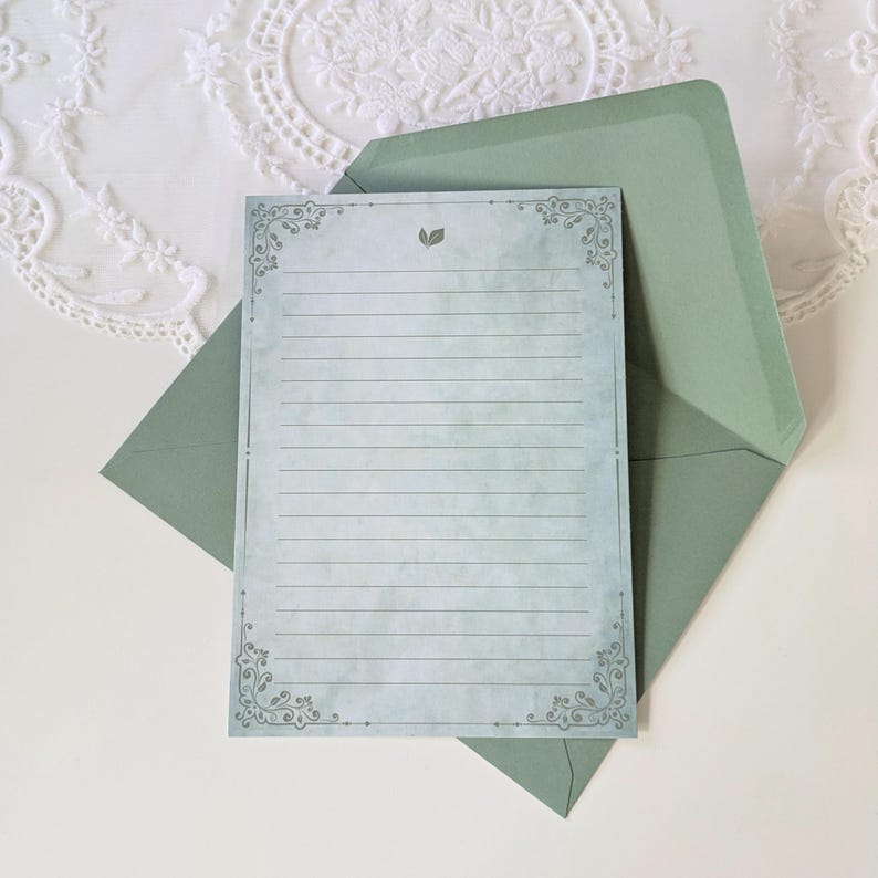 K&ouml;nnte beinhalten: Ein Briefpapier-Set mit einem linierten Schreibblatt mit dekorativen Eckdesigns und einer kleinen Blattillustration. Es liegt in einem salbeigr&uuml;nen Umschlag. Der Hintergrund ist eine wei&szlig;e Spitzentischdecke.