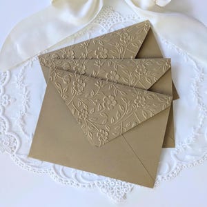 May include: Three gold-coloured envelopes with embossed floral designs. The envelope flaps feature a detailed pattern of flowers and leaves. The envelopes are arranged on a white lace doily and a white satin ribbon.
