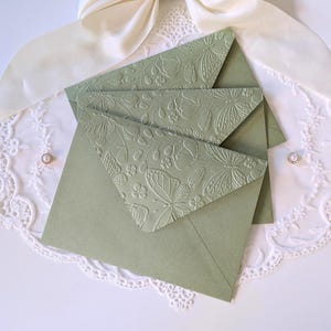 May include: Sage green envelopes with embossed butterfly and floral patterns. The envelopes are on a white lace surface, with a cream ribbon and a small earring.