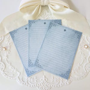 May include: Three pale blue lined writing paper sheets with decorative corner patterns and a seashell motif. The stationery is arranged on a white lace surface, with a cream-coloured bow in the background. Ideal for letter writing.