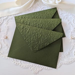 Pode incluir: Três envelopes verde-oliva com desenhos florais em relevo. Os envelopes estão dispostos em uma composição em camadas, com o envelope superior parcialmente aberto. O fundo apresenta uma toalha de renda branca e uma fita creme.