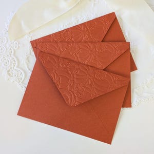 Puede incluir: Un conjunto de tres sobres de color terracota, con un diseño floral en relieve en las solapas. Los sobres están dispuestos sobre un fondo blanco de encaje y satén, creando un contraste de texturas y colores. Los sobres son probablemente para invitaciones o papelería.