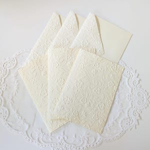 Peut inclure: Ensemble de papeterie ivoire avec motifs floraux en relief. L'ensemble comprend plusieurs cartes pliées et des enveloppes, disposées sur une nappe en dentelle blanche. Le motif en relief présente des fleurs et des vignes délicates, créant une apparence élégante et texturée.