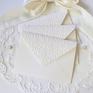 Può includere: Buste color avorio con motivi floreali in rilievo. Le buste sono disposte su una superficie di pizzo bianco, con un nastro color crema sullo sfondo. È visibile un ornamento decorativo di perle e strass.