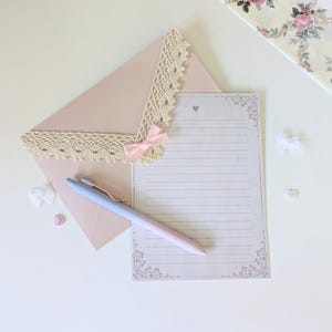 May include: A pink envelope with lace trim and a pink bow, a white lined writing paper with a decorative border, and a blue and pink pen.