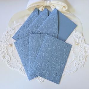 May include: A set of light blue stationery with embossed floral patterns. The set includes envelopes and cards, arranged on a white lace doily. A cream-colored bow and a small rhinestone embellishment are also visible.