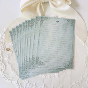 K&ouml;nnte beinhalten: Ein Stapel liniertes Briefpapier mit Vintage-&Auml;sthetik. Das Papier hat einen blassblau-gr&uuml;nen Farbton mit dekorativen Eckverzierungen und einem kleinen Blattdesign. Das Briefpapier ist auf einer wei&szlig;en Spitzentischdecke mit einer cremefarbenen Schleife angeordnet.
