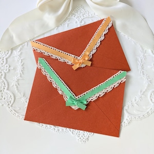 Könnte beinhalten: Zwei rostfarbene Umschläge mit dekorativer Spitzenborte und kleinen Schleifen. Ein Umschlag hat orangefarbene Verzierungen und eine Schleife, der andere grüne Verzierungen und eine Schleife. Die Umschläge liegen auf einer weißen Spitzendecke.