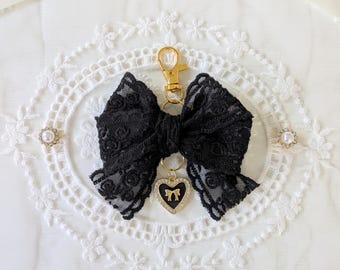 Peut inclure: Porte-clés en forme de nœud en dentelle noire avec un fermoir doré et un charm en forme de cœur. Le charm est noir avec un motif de cœur doré et entouré de petits strass transparents. Le nœud est sur un fond de dentelle blanche.