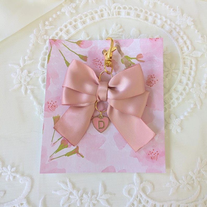 Peut inclure: Un porte-cl&eacute;s en forme de n&oelig;ud en satin rose clair avec un fermoir dor&eacute; et un charm en forme de c&oelig;ur. Le charm porte la lettre "D". Le n&oelig;ud est attach&eacute; &agrave; une carte florale rose. Le fond est en dentelle blanche.