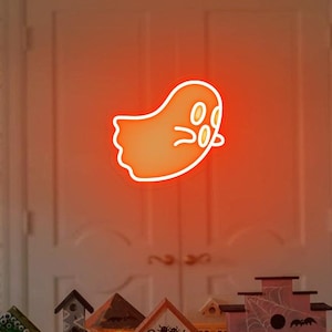May include: A neon orange ghost sign with a white outline and three white circles for eyes. The sign is hanging in front of a white door. There are several miniature houses with Halloween decorations in front of the door.