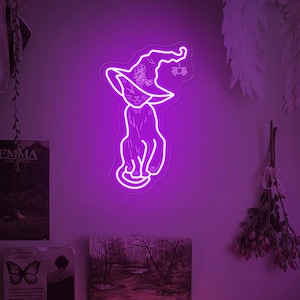 Könnte beinhalten: Ein lilafarbenes Neon-Schild mit einem Katzentier, das einen Hexenhut trägt. Die Katze sitzt mit dem Schwanz um die Beine gewickelt. Das Schild befindet sich an einer Wand mit anderen Dekorationen.