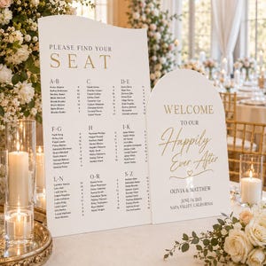 Könnte beinhalten: Zwei weiße Hochzeitsschilder mit goldfarbenen Buchstaben. Auf einem Schild steht "BITTE FINDEN SIE IHREN PLATZ" mit den Gästenamen in alphabetischer Reihenfolge. Das andere Schild lautet "WELCOME TO OUR Happily Ever After" mit den Namen des Paares und dem Hochzeitsdatum.