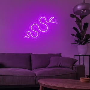 May include: A white neon snake sign with a coiled body and a raised head, illuminated in a bright purple light against a purple wall. The sign is mounted above a purple couch with a wooden coffee table in front of it.