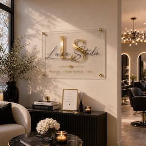 Może przedstawiać: Przezroczysty akrylowy szyld ścienny ze złotym napisem LS Luxe Studio oraz tekstem BEAUTY CONFIDENCE YOU. Szyld jest zamontowany za pomocą czterech złotych dystansów na beżowej ścianie nad czarną konsolą z elementami dekoracyjnymi.