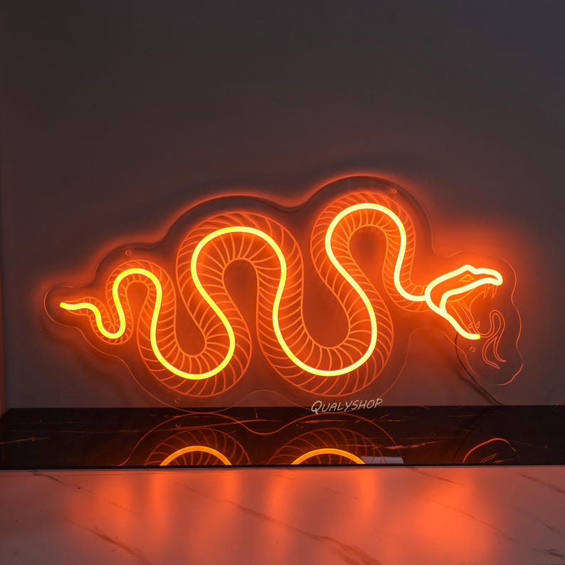 May include: Orange neon light shaped like a snake, with a white background. The neon light is on and glowing. The light is mounted on a wall.
