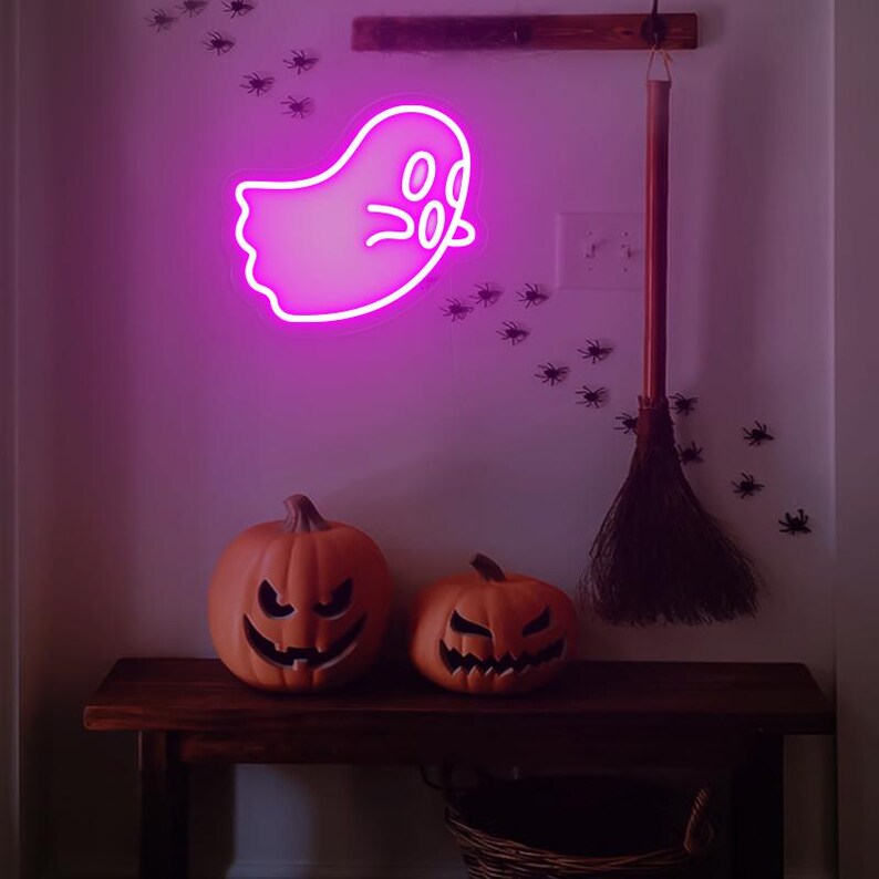 May include: A pink neon ghost sign with a cute face, hanging on a white wall with a broom and black spider decorations. Two orange pumpkins with carved faces sit on a wooden table.