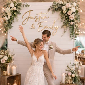 May include: A white shiplap backdrop featuring gold script lettering that reads Justin & Emily. The display is adorned with large floral arrangements of white and blush roses with eucalyptus greenery, positioned on both top corners of the wall.