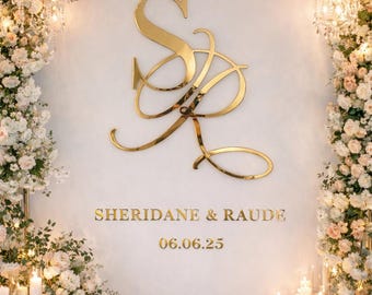 May include: A large gold metallic monogram featuring the letters S and R in a cursive script, positioned above the text SHERIDANE & RAUDE and the date 06.06.25, all displayed on a plain white backdrop framed by white and pink floral arrangements.