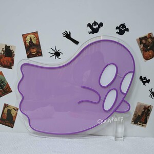 May include: Purple neon ghost sign with a white outline. The sign is shaped like a ghost with two eyes and a mouth. The sign is on a white background with other Halloween decorations.