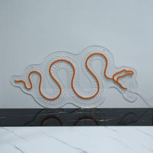 May include: Orange neon snake design on a white wall. The snake is outlined in white and has a textured pattern.