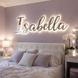 May include: A wall-mounted, reflective silver sign displaying the name Isabella in a cursive script font. The sign features integrated backlighting that casts a soft, warm glow against the wall, creating a halo effect around the letters.