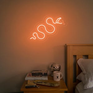 May include: A white neon snake sign with an orange glow, mounted on a wall above a wooden nightstand with books and a mug.
