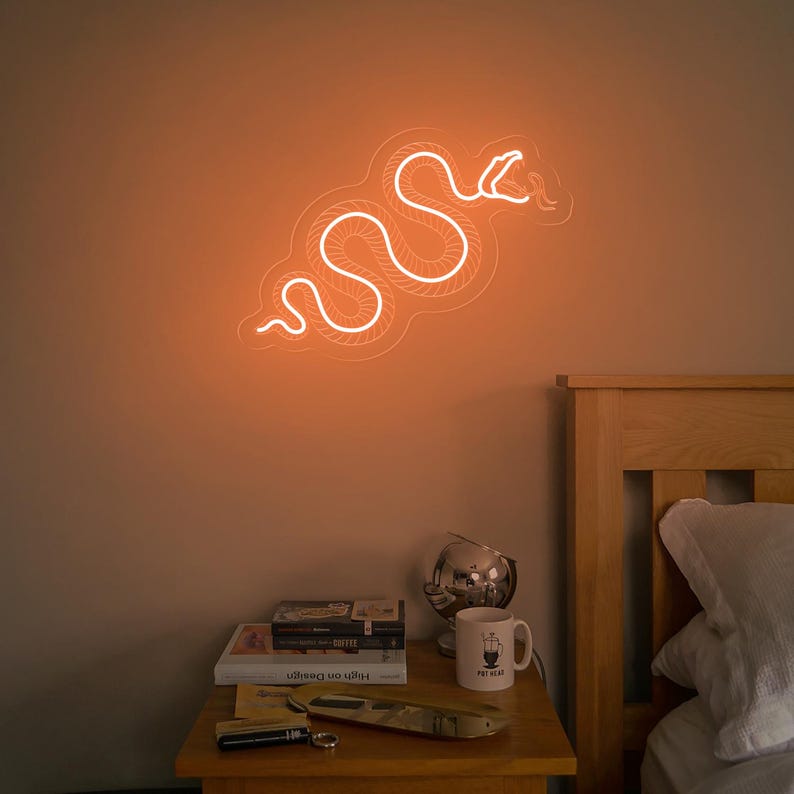 May include: A white neon snake sign with an orange glow, mounted on a wall above a wooden nightstand with books and a mug.