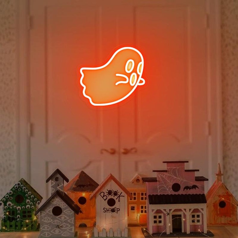 May include: A neon orange ghost sign with a white outline and three white circles for eyes. The sign is hanging in front of a white door. There are several miniature houses with Halloween decorations in front of the door.