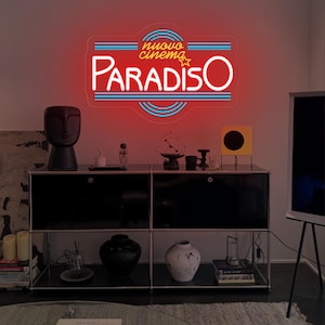 Puede incluir: Un letrero de neón con las palabras "Nuovo Cinema Paradiso" en rojo, blanco y azul. El letrero está montado en una pared blanca sobre un mueble negro. El tema general es el cine y la decoración del hogar.