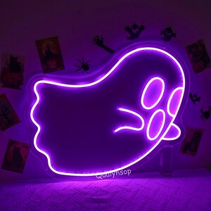 May include: Purple neon ghost sign with a white outline. The ghost has two eyes and a mouth. The sign is lit up and casting a purple glow on the wall behind it.