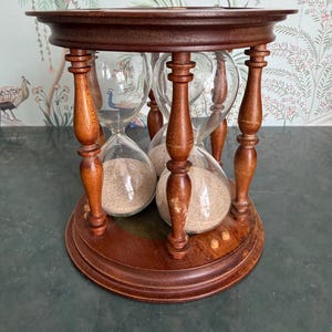 May include: A wooden hourglass with four glass chambers filled with sand. The hourglass is supported by a dark brown, turned-wood frame with a round base and top. The glass chambers are clear, and the sand is a light tan color.