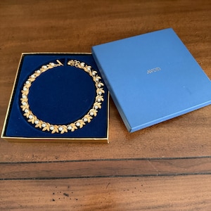 May include: A gold-toned necklace featuring a repeating leaf pattern with small white faux pearls, presented in a square blue box with the text AVON printed in gold on the lid. The necklace is displayed inside the box on a dark blue velvet insert.